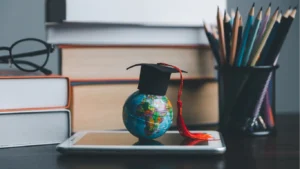 Miniature Globe sitting atop an iPad in a school desk environment with a graduation hat on top, illustrating the concept of a Global School. The depiction is enhanced by background textbooks and pencils. The iPad further alludes to the Global School concept of connectedness and the online landscape.